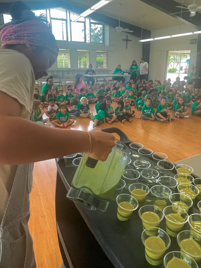 a woman pouring a juice blend into cup in front of an audience of children