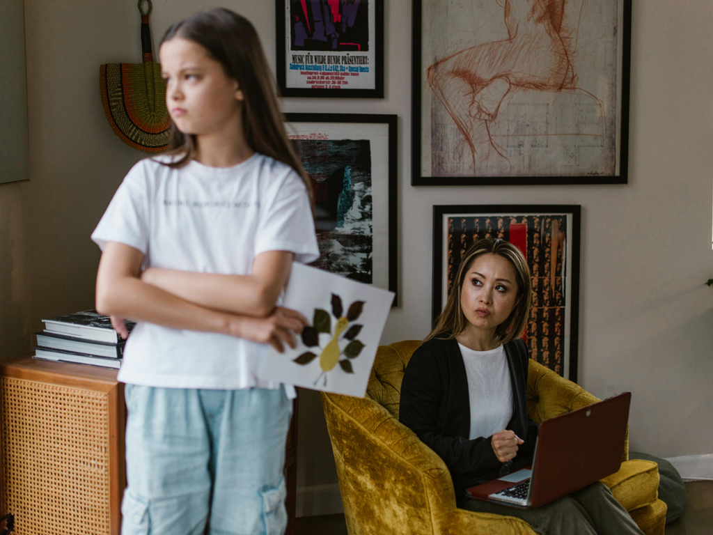 A girl stands with folded arms while a woman sitting in a yellow chair with a laptop looks at her.