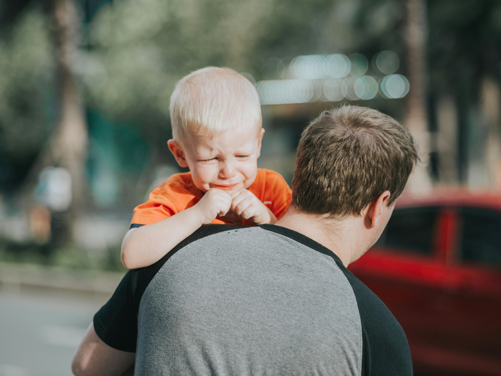 A young child crying while being carried on the back of an adult outdoors near a street with a red car in the background.