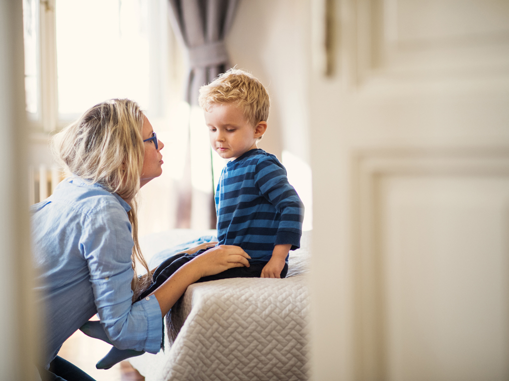 A young mother talking to her sad toddler son inside in a bedroom.